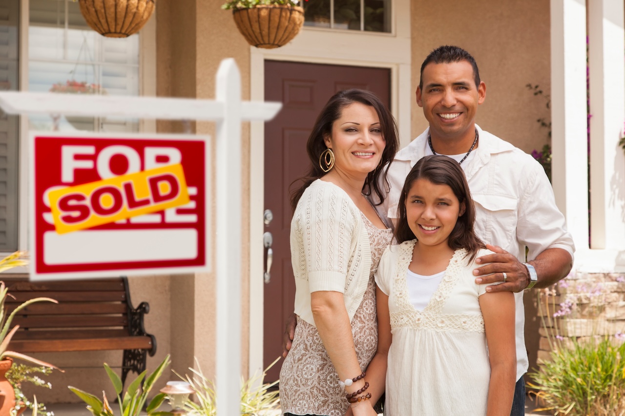 Madre, padre e hija hispanos delante de su nueva casa con el cartel de Se Vende Casa Inmobiliaria.