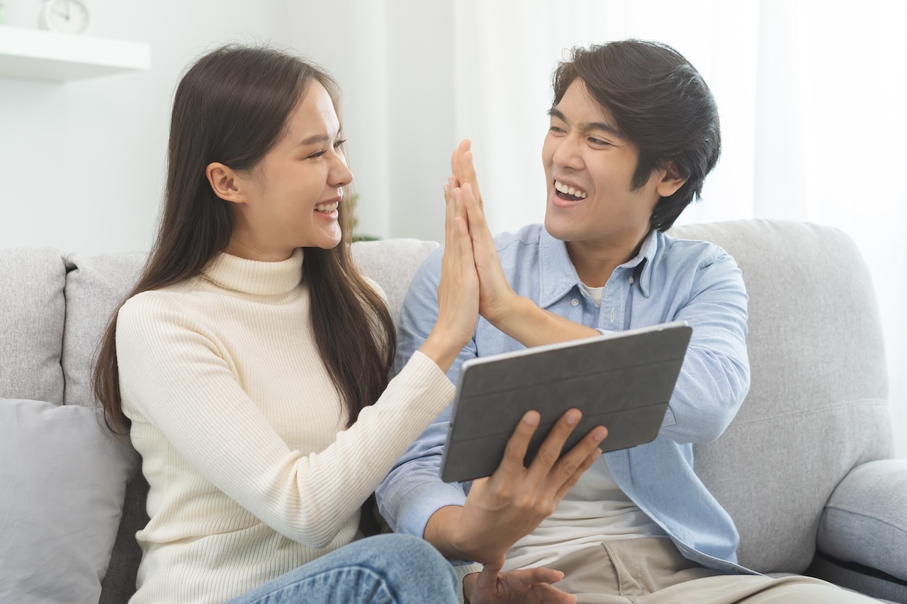 Happy Excited, Smiling Asian Young Couple Love Using Tablet Pc, Great Deal Or Business Success, Received Or Getting Cash Back, Tax Refund, Good News By Mail While Sitting On Sofa, Couch At Home.