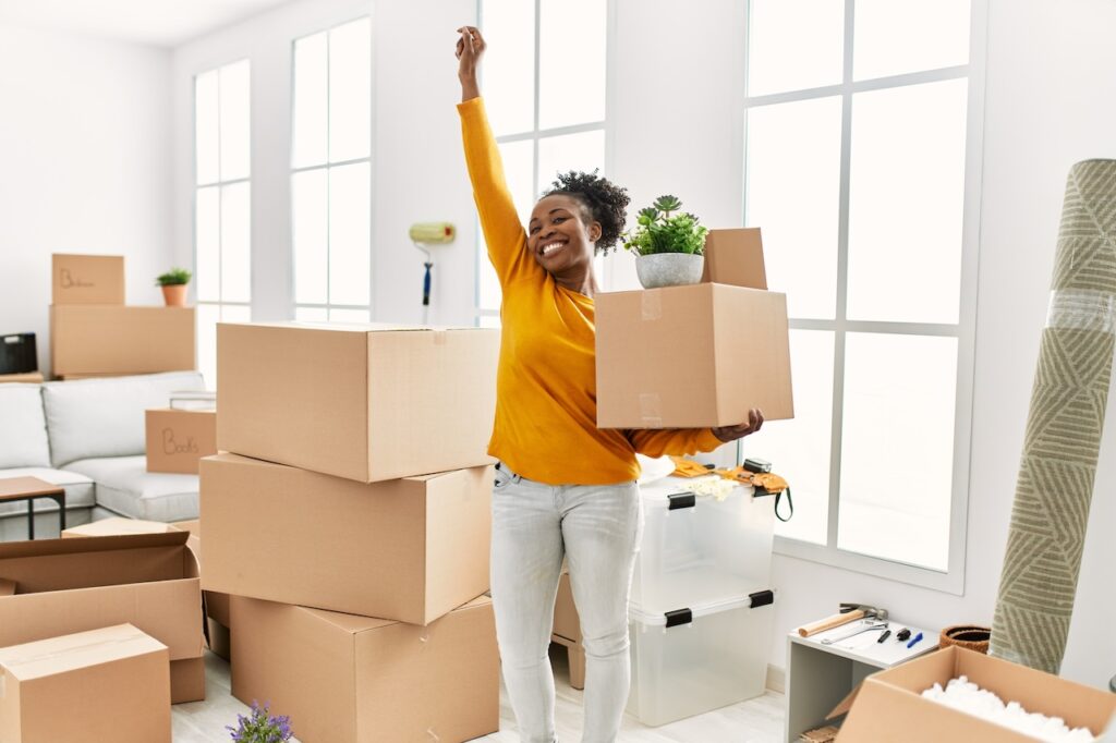 African American Woman Holding Package Standing With Winner Expression At New Home