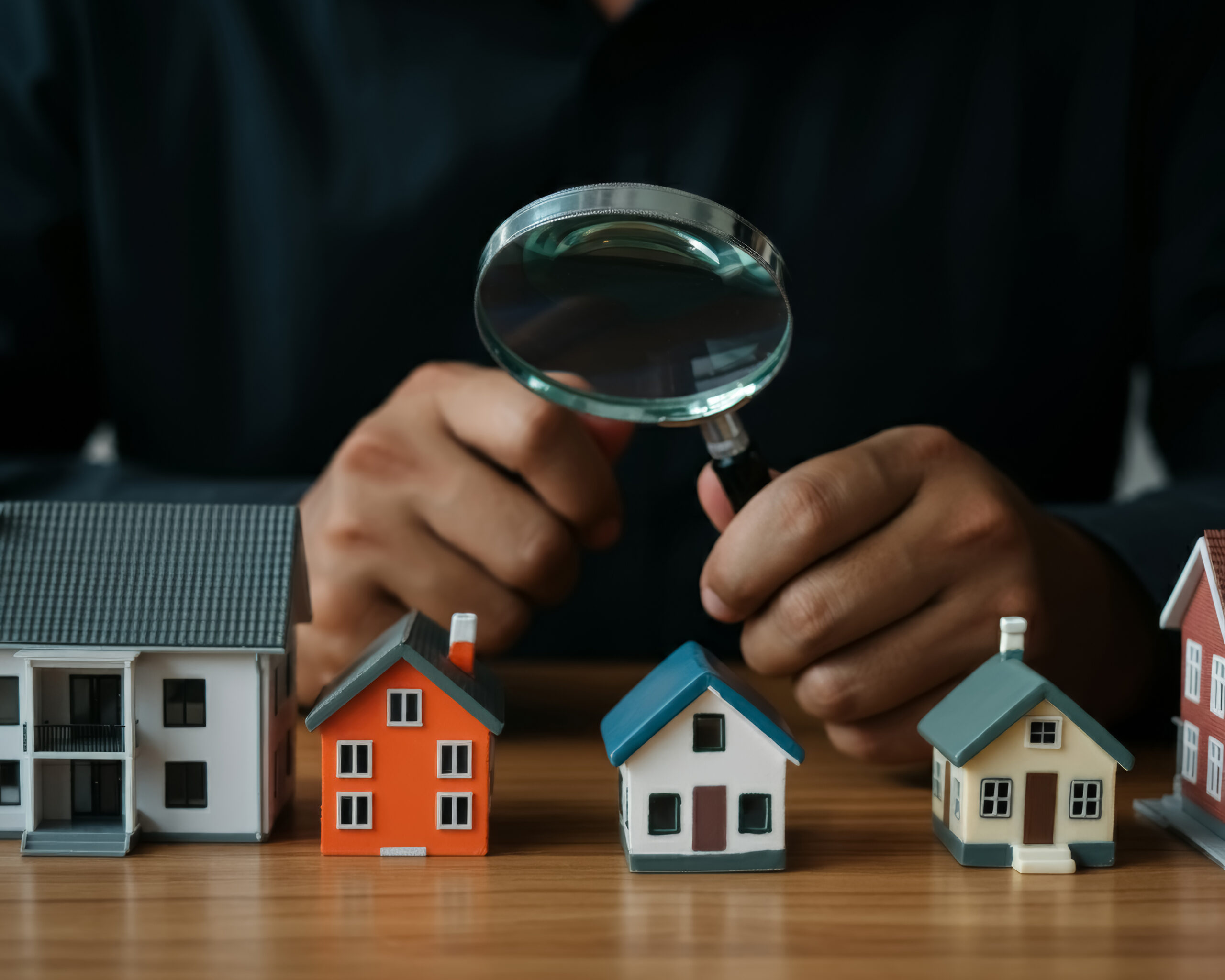 Man's Hand Holding Magnifying Glass And Looking At Tiny House Model, House Selection, Real Estate Concept.